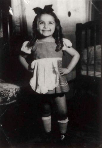 Black and white photo of a young girl smiling and standing indoors, wearing a checkered dress with a bow in their hair and knee-high socks.
