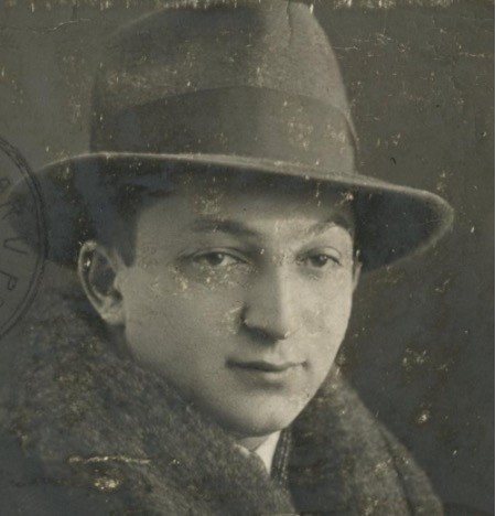 Close-up sepia photograph of a man wearing a hat and a fur-collared coat. The expression is serious, and there's a slight blemish on the face possibly from the age of the photo. A postal stamp partially visible on the top left corner, suggesting the photo might be from an old passport or document.
