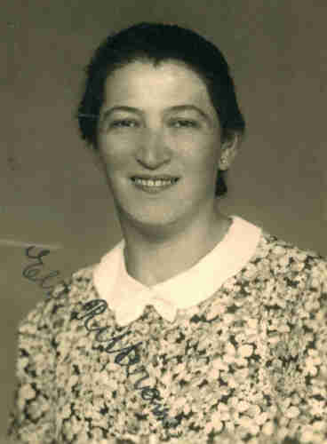 Portrait ID photo of a woman. She has a blouse with flowered pattern. She has dark hair pinned in the back. She is smiling exposing her upper teeth. She is looking into the camera.