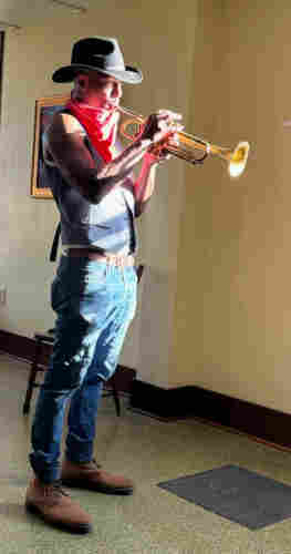 Man in a black cowboy hat and vest, standing in streaming sunlight, playing a trumpet 