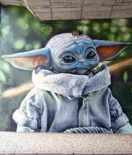 Streetartwall. A fantastic mural of a baby Yoda (Star Wars) in "photorealistic" style was sprayed on the concrete wall under a bridge. The gray, cuddly alien is depicted halfway up. He is wearing a thick winter jacket with a high fur collar and is standing in front of a green landscape.
Info: A baby Yoda is a green/grey creature between animal and human, has long ears and big eyes and is very cute. It's a figure from the Star Wars Universum.