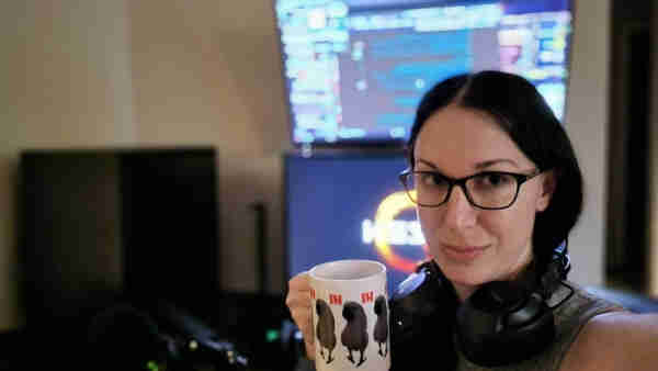 Selfie of a woman with black hair and black glasses sitting in front of a computer setup holding a white coffee mug depicting a Moluccan cockatoo saying "Hi."