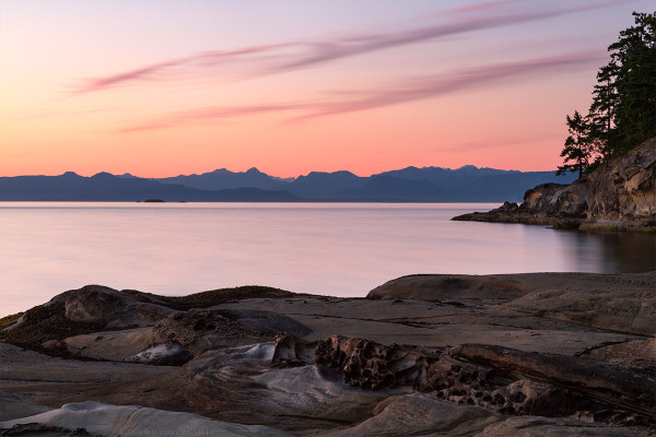 Two lines of clouds and the sky over distant mountains show the orange glow of sunset with a sandstone shoreline and ocean in the foreground.