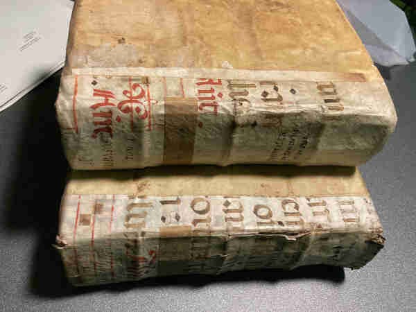 Two weathered and aged books with text in a Latin script on their spines, lying on a dark surface, with some papers in the background.