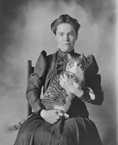 Black and white photo of a white woman in a dark dress or blouse and long skirt, sitting up in a chair for a studio portrait and cradling a shorthaired tuxedo tabby cat in her arms. Both of them look straight at the viewer.