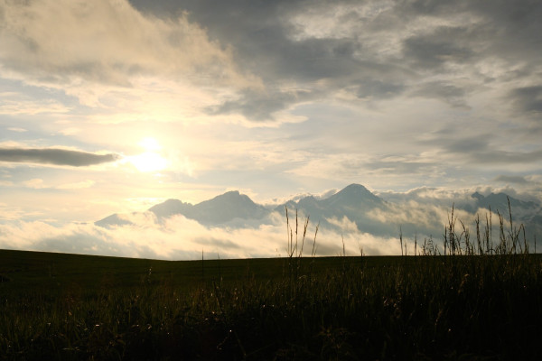Mountains with clouds hovering around. Sun in between of clouds above mountains. Dark green field, with blades of grass, in foreground of photo.