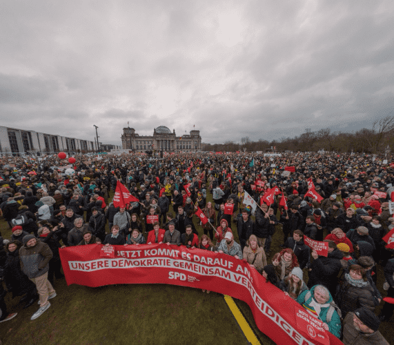 Im Vordergrund ein rotes Banner der SPD, auf dem steht: Jetzt kommt es darauf an: Unsere Demokratie gemeinsam verteidigen. Dahinter tausende Menschen, die vor dem Reichstag demonstrieren.