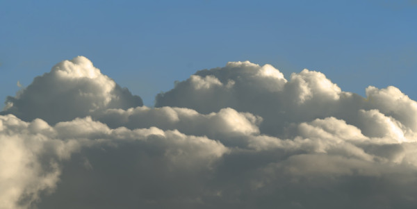A zoomed-in shot of some horizontal fluffy clouds just above the horizon, lit by a low sun, creating dramatic dark and bright parts of the clouds. The sky above is a pale blue and the button of the clouds are a dark grey color.