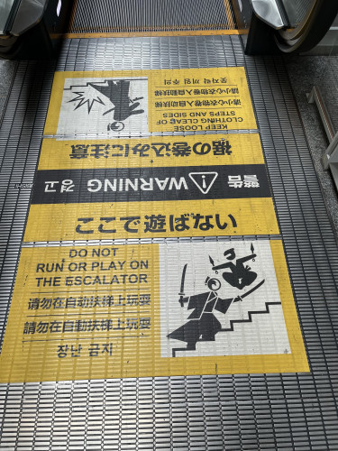 Warning signs at the base of an escalator with instructions in multiple languages advising not to run or play on the escalator and to keep loose clothing clear of steps and sides.