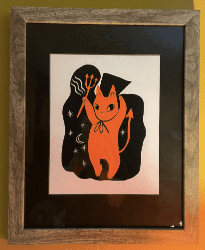 A print in a grey wooden frame with black mat board against a mustard yellow wall. The print depicts an adorable cartoonish anthropomorphic kitty in a classic red-orange devil costume, waving a little pitchfork in the air.