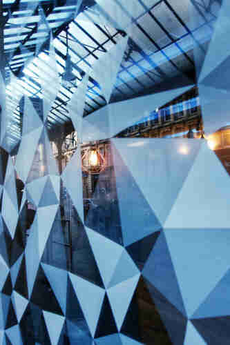 A geometric patterned glass wall with reflections of an indoor space with hanging lights and an exterior view of a building.