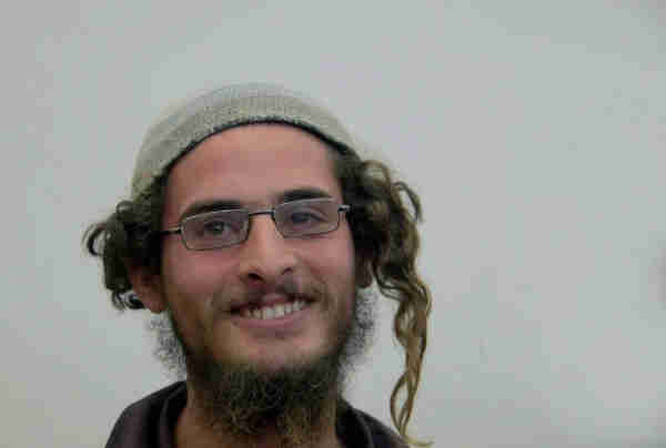 A smiling man with glasses, curly hair, and a beard wearing a knitted cap on a plain background. Meir Ettinger, Meir Kahane’s grandson and leading ideologue of the hilltop youth