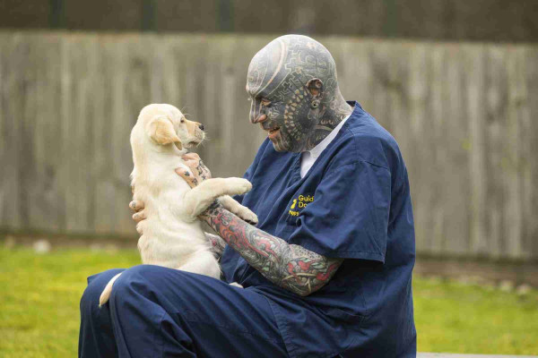 A man with numerous blue coloured tattoos (covering his head and arms, all visible body parts) sits wearing Guide Dogs branded navy blue scrubs. The man is holding a young yellow Labrador guide dog puppy in his lap. There is a grassy area and fence in the background.
