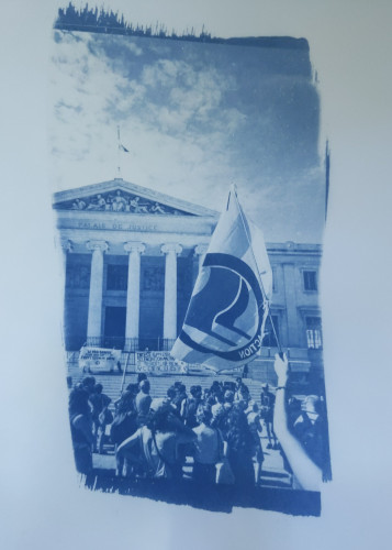 Tirage cyanotype en bleu et blanc.
Photo avec en fond le palais de justice de Marseille avec des colonnes, sur les marche devant des manifestants et manifestantes tenant des banderole, encore devant il y a la manifestation de dos et au premier plan un bras sort du coin du bas a droite en brandissant un drapeau trans antifa