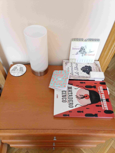 Photo of a bedside table with a coaster with a drawing of a Madrid on it. A lamp that's activated my touch and not a button, an art calendar drawn by Rike, a yearly planner that says Yay for 2024, the graphic novel Slaughterhouse Five and the paperback The Book Eaters and a box with earplugs