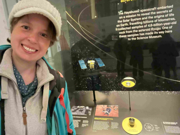 A selfie of me standing in front of the display case at the science museum containing the asteroid grain. The grain is in a small metal cylinder in a yellow circle at the bottom of the display case. A mirror reflects the contents of the cylinder for better viewing. 