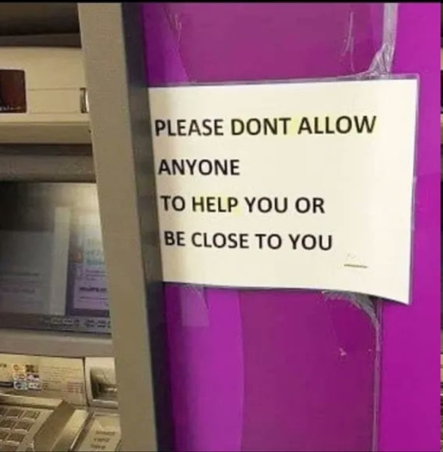 Still image. Cash machine with a bright purple/pink wall to its right. There is a laminated sign pasted up, black print on white paper, reads:
"PLEASE DONT ALLOW
ANYONE
TO HELP YOU OR
BE CLOSE TO YOU"