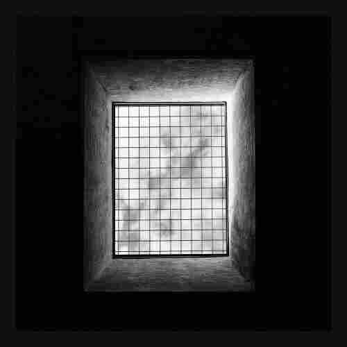 This black and white image shows a rectangular window with a grid pattern, looking out to a cloudy sky. The window is deeply recessed into a rough-textured wall, creating a sense of depth. The contrast between the bright outside light and the dark interior emphasizes the grid's lines, giving the image a symmetrical and geometric appearance. The overall effect is minimalist, highlighting the interplay between light and shadow.