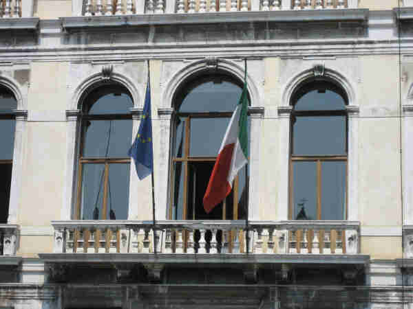 A photo showing three arched windows on the façade of a classical building with a white and weathered exterior. Two flags are hanging from the central window's balcony: the European Union flag on the left and the Italian flag on the right. The balcony features a stone balustrade and the architecture show a historical European setting.