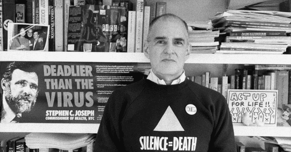 a photograph of Larry Kramer in his library. he is wearing a sweatshirt with Act Up's lavender triangle and underneath it their catchphrase: SILENCE = DEATH

behind him is a poster with the face of the then NYC Health  Commissioner, Stephen C. Joseph. it says: Deadlier than the virus.

