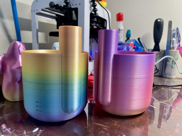 Two pastel ombre PLA printed reservoirs for self-watering pots sitting on a brown wood desk; each has a round tube on the side to deliver water. The pot on the left is golden yellow to green to purple. The pot on the right is purple to pink to peach in color. A small printer and various tools are visible in the background. 