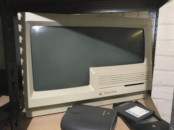 A photo of a wide Classic look Macintosh labeled SE/30L with a strange inverted L-display. It has a single floppy drive at the front, and the giant display is also a strange bubbled. It sits on a shelf with a QuickTake 150, Newton, and a Mac install zip disk.