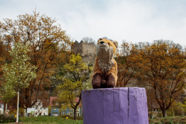 Red fox plushy sitting on a tree stump that has been painted purple. In the background are a couple trees and castle ruins on top of a cliff.