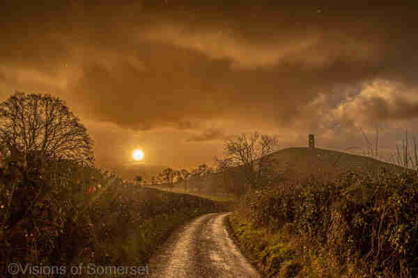 Golden sun to the left of the scene. straight ahead is the Tor and tower. A very golden scene but with raindrops visible. 