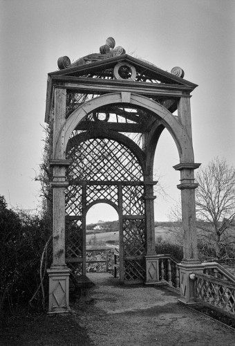 An ornate wooden structure stands at the end of a terrace. Banisters can be seen on the lower right, suggesting steps going down to a lower level. Through the window at the far end part of the castle wall is visible and beyond it a white house in some woodland. Portrait format photo in black and white.