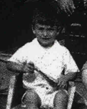 Blurred photograph of a young boy sitting on a garden chair. He is wearing clothes with short sleeves and legs. He is holding something in his hands. 