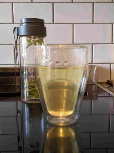 Freshly poured green tea in a double walled borosilicate glass, with a glass tea flask in the background, filled with spent green leaves. 

The two vessels are pictured on top of a glass induction cooktop.