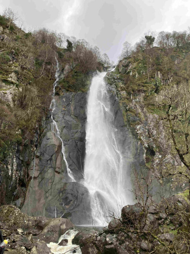 Water from Aber Falls, in Coedydd Aber National Nature Reserve, cascading down a rocky cliff surrounded by vegetation. A dark grey sky above.