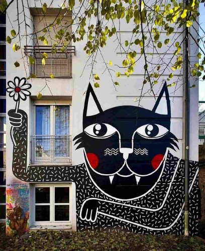 Streetartwall. On the outside wall of a multi-storey building is a large mural of a black cat sprayed/painted in a naive pop art style. The head is large, with big eyes, two long teeth, red cheeks, waves for whiskers and two pointed ears. It has black fur with small white strokes in all directions. The black and red cat is holding a small white flower in its hand and looks very friendly.
Info:
Joachim is a young highly talented street artist from Antwerp, Belgium. His distinctive graffiti pop style coupled with a wide range of styles is unmistakable. He also brings intricate textures and wild color compositions to canvas when working in his own studio. Meanwhile, art galleries have also discovered him and if I may note and...he is a favorite artist of mine.