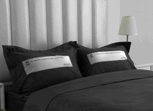 A photo of a black bed with black pillows, and across each pillow is the final macos dialog saying "It is now safe to switch off your consciousness", instead of your computer.