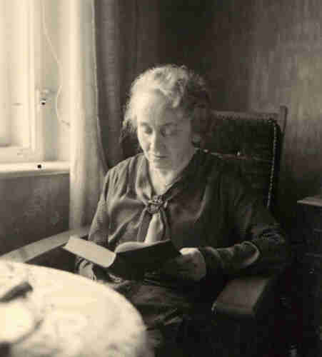 An elderly woman sits in an armchair by the window. She is reading a book. In front of her is a table with a white tablecloth.