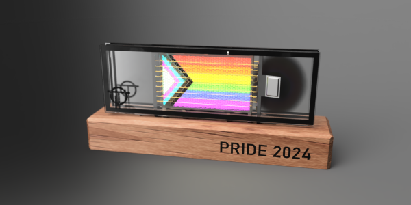 Rendering of a flat panel style vacuum fluorescent display. The anode the progress pride flag. The panel is mounted on a wood block with the words "PRIDE 2024" engraved on the front. The cathode wire glows dimly and the anode behind it can be seen glowing brightly through the hexagonal grid.