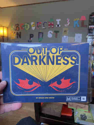 A hand holding a book titled "Out of Darkness" by Micah and Snow, with a retro sci-fi design featuring red eyes against a blue background. The book is for use with the Mothership Sci-Fi Horror RPG.