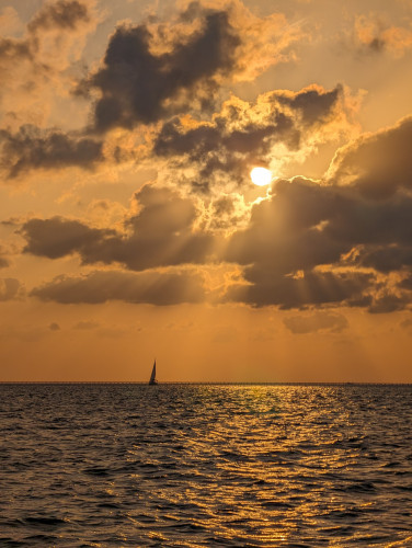 The sun is peeking through dark clouds over even darker water. Its light is orange and its rays are visible where they pierce the clouds. A lone sailboat is silhouetted against the horizon.