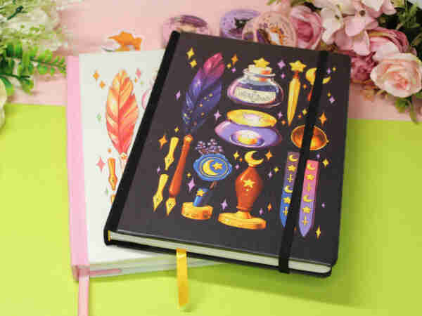 Two notebooks stacked on top of each other: top one is black with purple/gold stationery drawings such as feather quill, ink bottle, wax seal and letter opener. bottom one is mostly obscured and is minty green with pink/yellow feather quill and colorful sparkles. 

Backdrop is green/pink with some fake roses and some washi tape/stickers