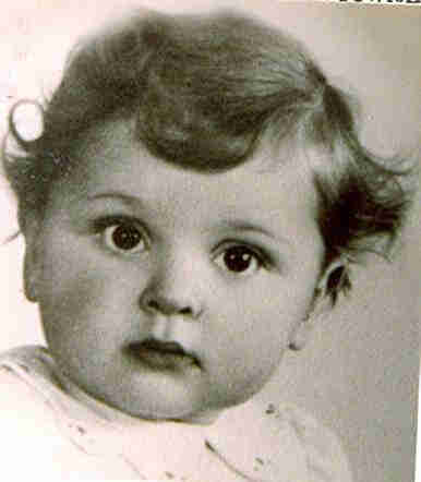 Picture of a little girl's face. She has slightly wavy hair. She has a wide round face, large dark eyes looking into objects. She is wearing a white blouse with some dark dots.