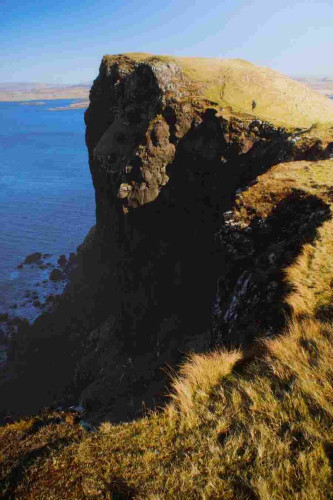 This image shows a rugged seascape with a steep, craggy cliff high above the sea loch. The cliff is covered in yellow grass. There is a small person hiking up towards the top of the next cliff, providing a sense of scale to the scene. The water below is calm, and the sky is clear with a gradient of blue hues.