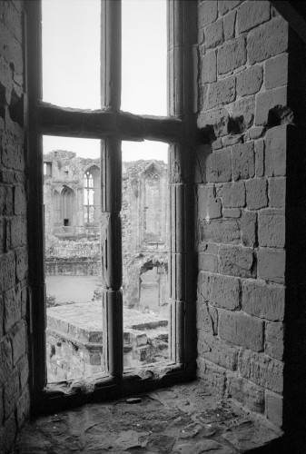 Black and white portrait format photo of a tall window with stone frame set i a thick stone wall. The window looks out on a ruined castle landscape. It is from the building Robert Dudley built for Elizabeth 1's last visit.