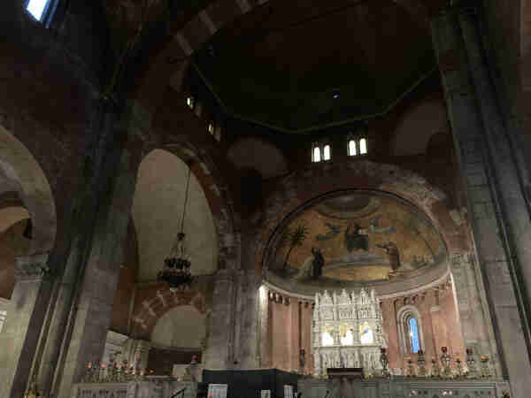 Interior of a cathedral showing altar, apse with frescoes, Gothic architecture, and hanging chandelier.