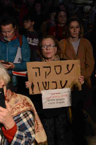 An elderly woman in a crowd holding a sign that reads “Deal Now!” A piece of paper is attached to it reading: “359,000 destroyed houses in Gaza. Stop destroying. Start talking!”.