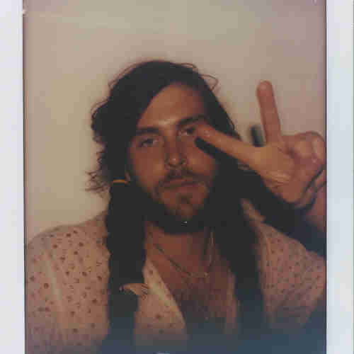 Polaroid-style photograph of a man giving a peace sign with his hand. He has long, dark hair with a portion braided on one side and secured with a feather. He wears a light-colored shirt with a subtle floral print and a visible necklace, conveying a bohemian or “hippie” style. His expression is relaxed and friendly, and the overall vibe of the photo is casual and laid-back. The image captures a moment that feels both personal and evocative of a certain carefree, retro aesthetic.