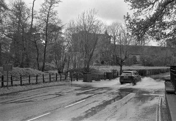 A car drives through water streaming over the road. Behind are trees, behind them the Castle. Black and white photo.