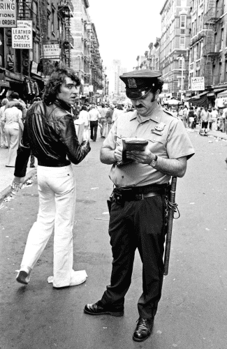 A policeman writes a ticket on a New York street as a man seemingly walks past or away from him, turning back to look at the camera.