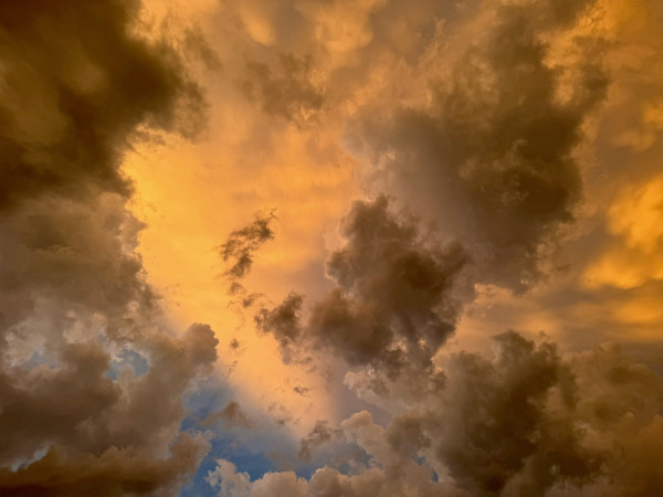 Quite dramatic, horizontal orientation, photo of the sky at dusk the clouds in the center are a golden orange color however some of the orange is blocked by various earing, dark clouds. Additionally vibrant, blue sky pokes through on the lower half of the frame along with sparse puffy white clouds. 