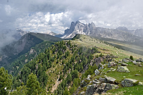 A photograph of a rugged mountainous landscape. In the foreground is a mountain ridge with sparse forests on the left and meadows on the right, both with large rocks in between. In the background are high, jagged peaks of higher mountains with no vegetation. The image is framed by low clouds on the left and right edge, and higher clouds at the top.