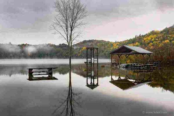 Structurs and a picnic table reflected on flood water, with distant hills in fog and fall color